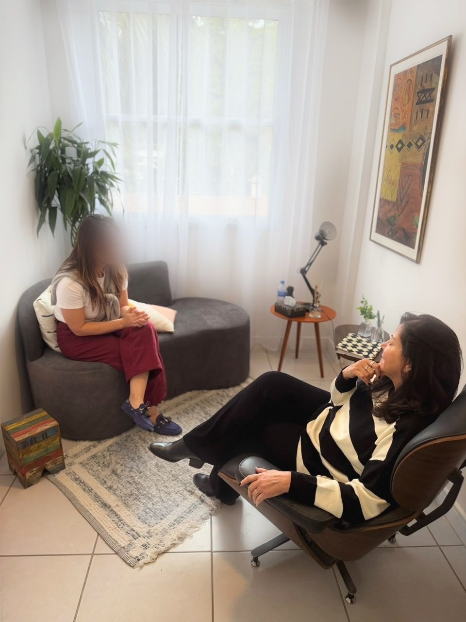 Consultório com sofá cinza, planta, tapete claro, quadro colorido na parede e luz natural entrando pela janela
